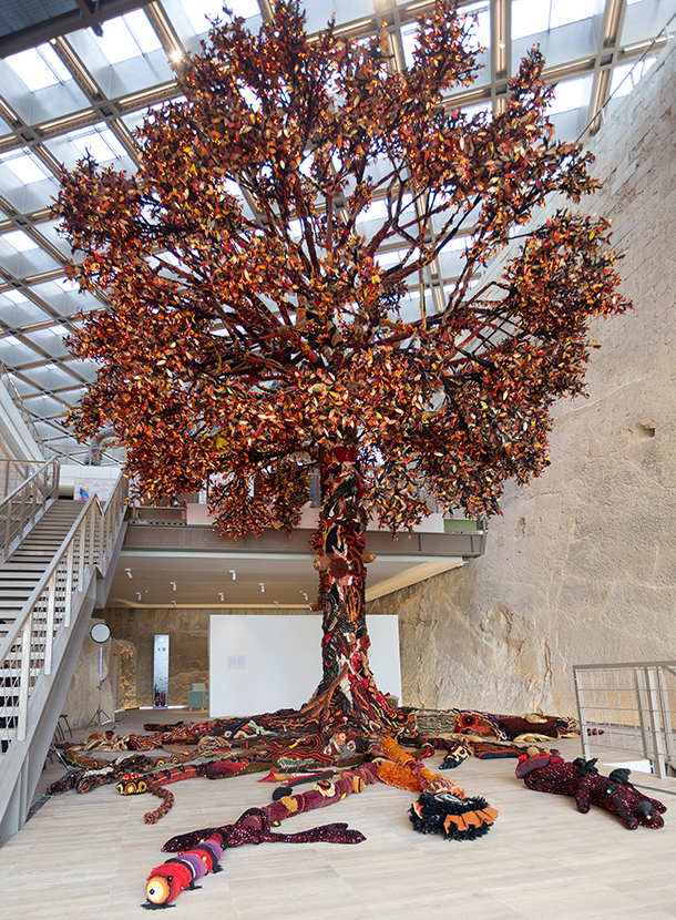 A large, colorful sculpture of a tree with roots and leaves made of fabric, displayed indoors.